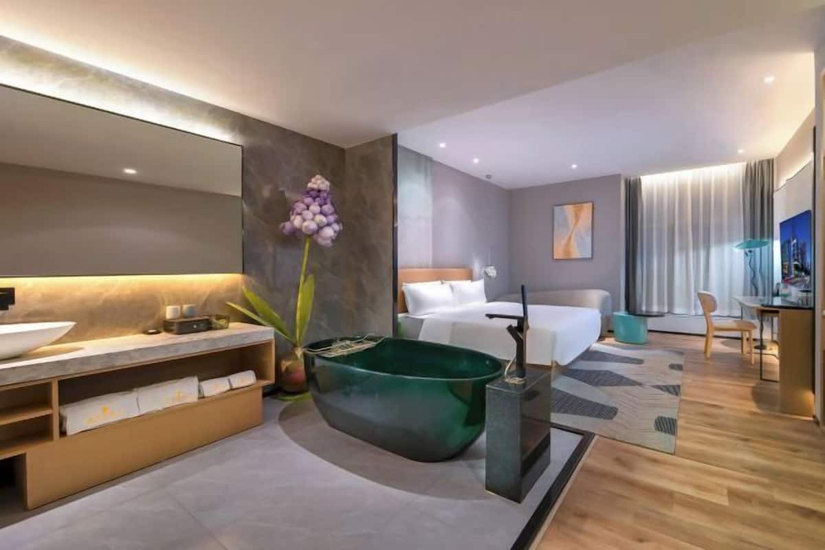 Ein modernes Hotelzimmer mit einem großen Bett, einer Sitzecke mit Tisch und Stühlen, einem großen Spiegel und einer grünen Vase mit Blumen.