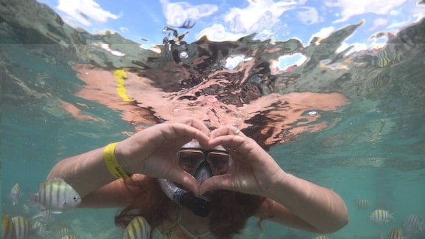 Buceador formando una forma de corazón con las manos bajo el agua, rodeado de peces.