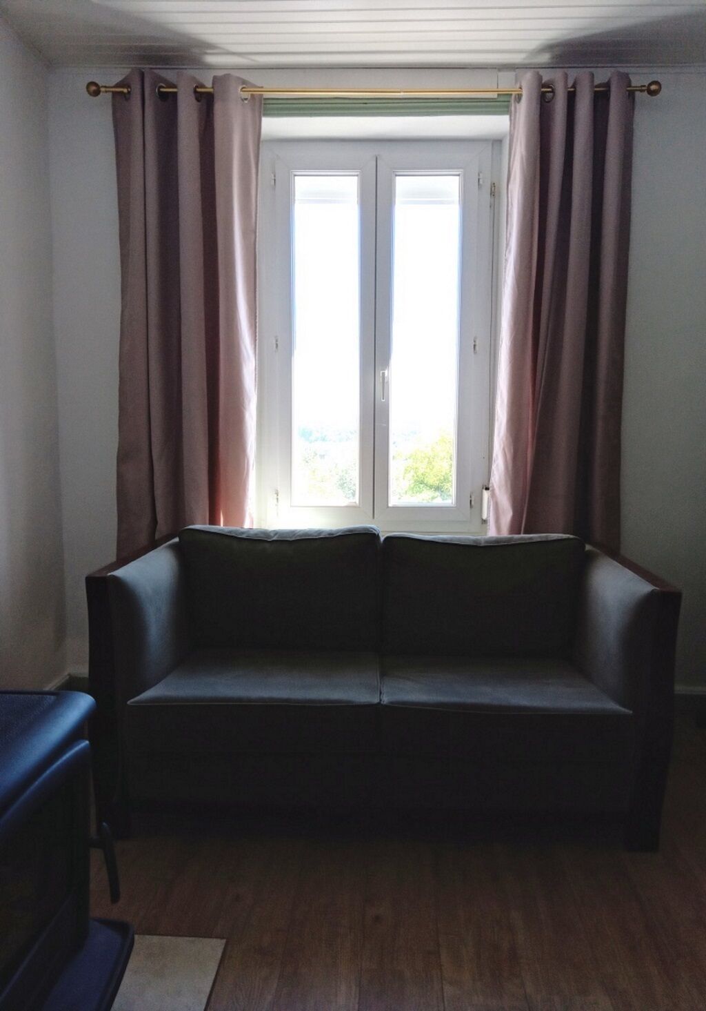 Una habitación con un sofá de color oscuro, piso de madera y una ventana con cortinas.