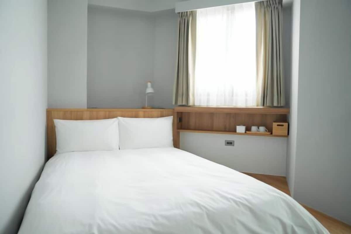 A hotel room with a bed, a nightstand, a lamp, a window with curtains, and a shelf with items.