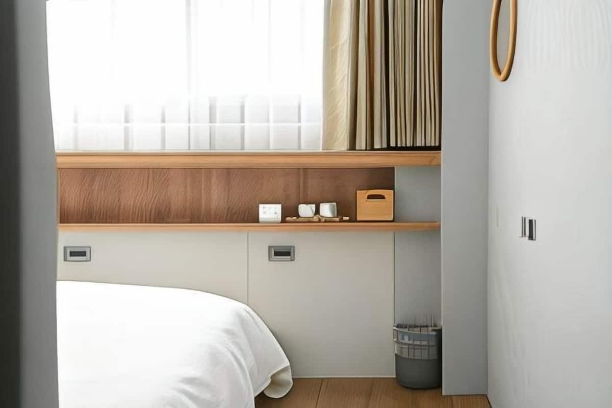 A bedroom with a bed, a window with curtains, a wooden shelf with items, and a small trash bin.