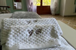 Una toalla blanca doblada con delicadeza y con iniciales bordadas sobre una cama en una habitación con suelo de baldosas y una ventana con cortinas.