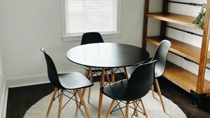 Un comedor con una mesa redonda, cuatro sillas, un tapete circular, una estantería de madera, un reloj de pared y una ventana con persianas.