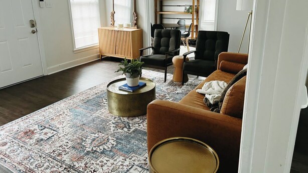 Una sala de estar con un sofá marrón, un sillón negro, un aparador de madera y una alfombra estampada.