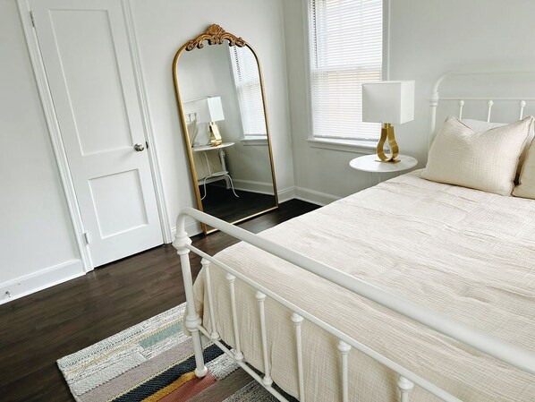 A bedroom with a white bed, a white door, a mirror, a bedside table with a lamp, and a window with blinds. - Welcome to your Memphis retreat steps from the vibrant University of Memphis! (Memphis)