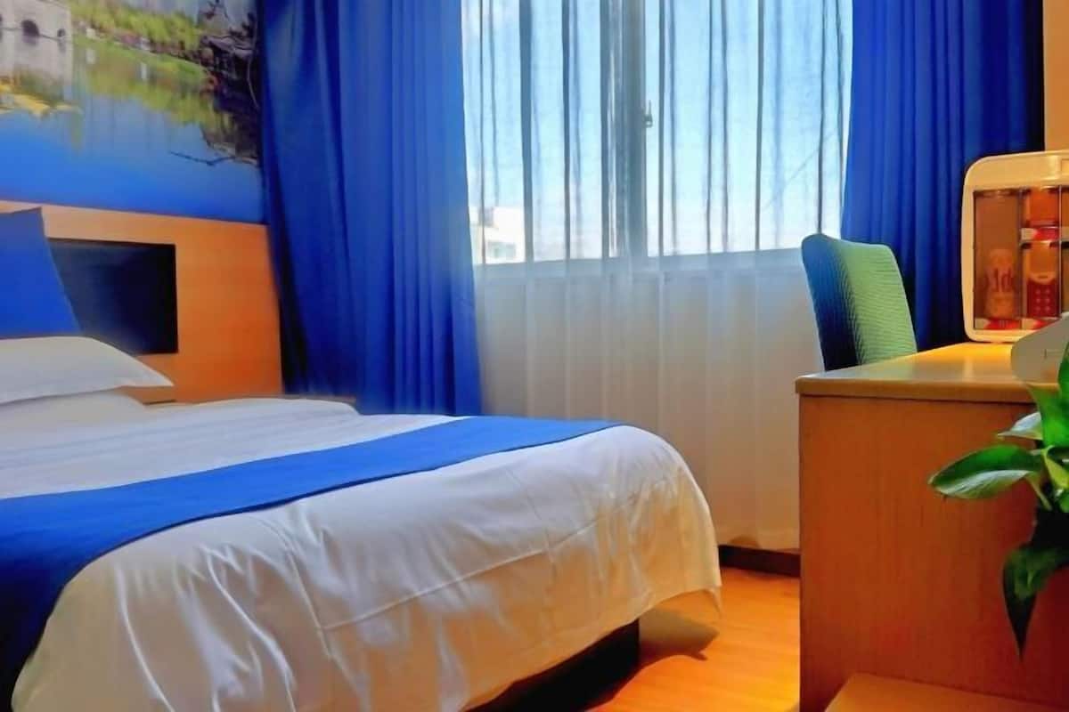 A hotel room with a bed, a desk with a lamp, a window with curtains, and a painting on the wall.