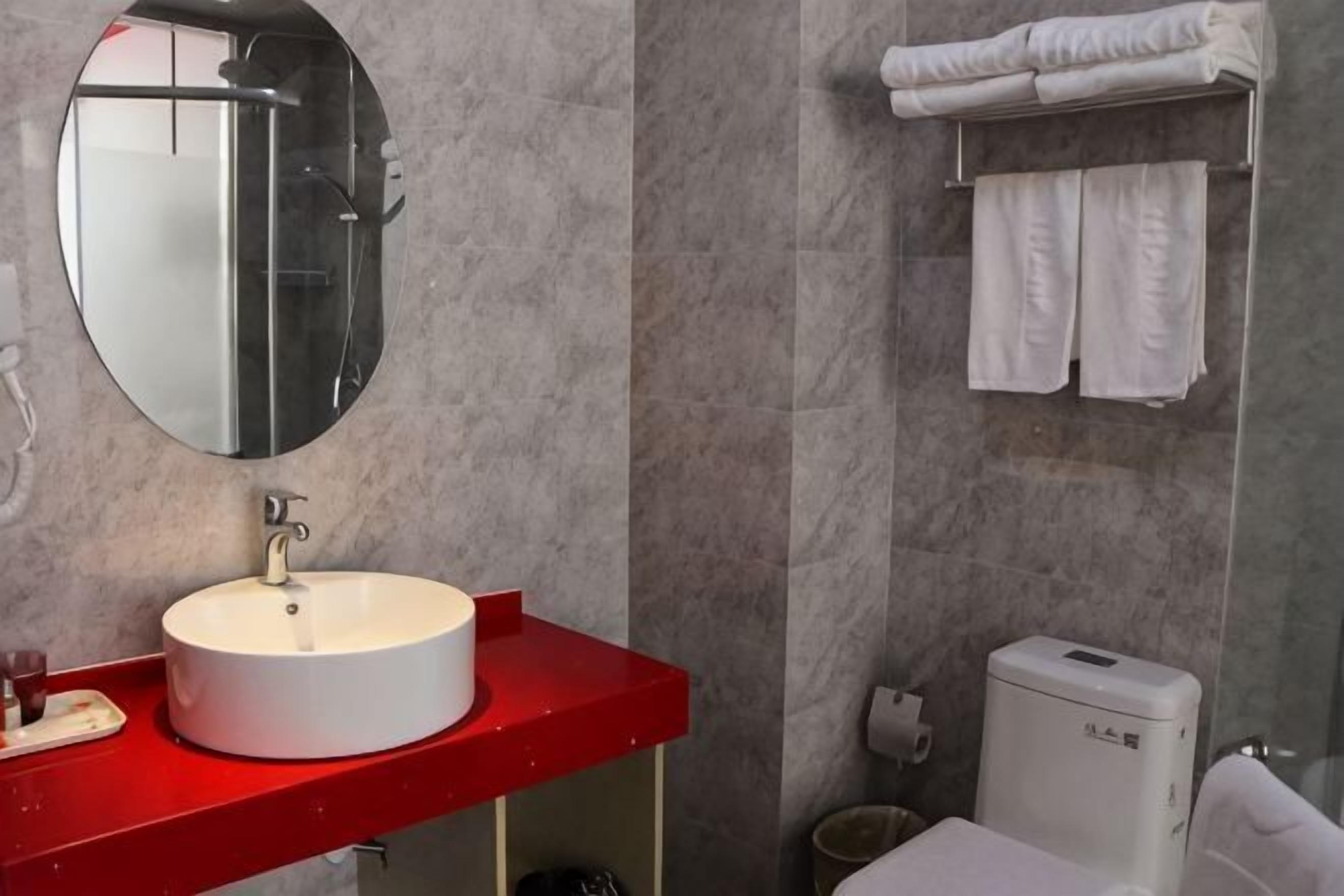 A bathroom with a round mirror, a white sink, a red countertop, a toilet, and a towel rack.
