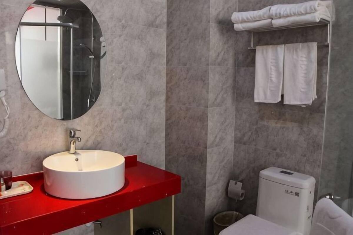 A bathroom with a round mirror, a white sink, a red countertop, a toilet, and a towel rack.