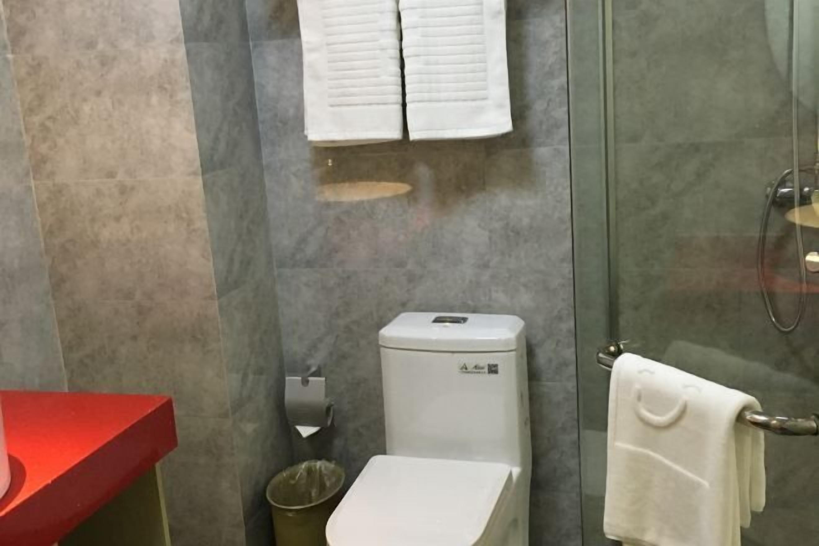 Un baño con cabina de ducha de vidrio, inodoro, lavabo rojo y toallero con dos toallas.