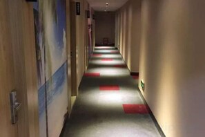 Un corridoio d'hotel con pavimento rivestito di moquette, porte sul lato sinistro e una parete con un poster.