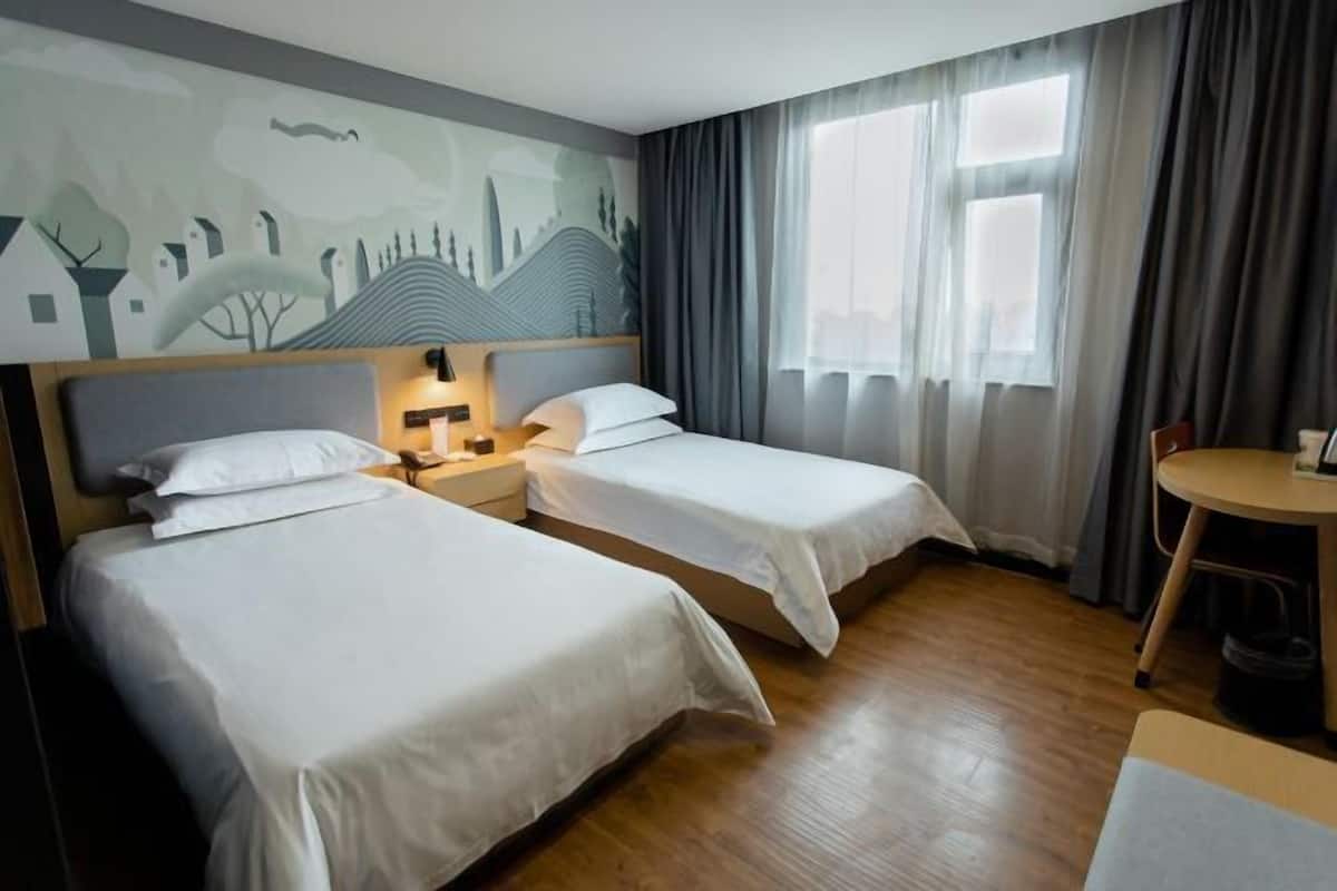 A hotel room with two beds, a nightstand with a lamp, a small table, and a view of a cityscape mural on the wall.