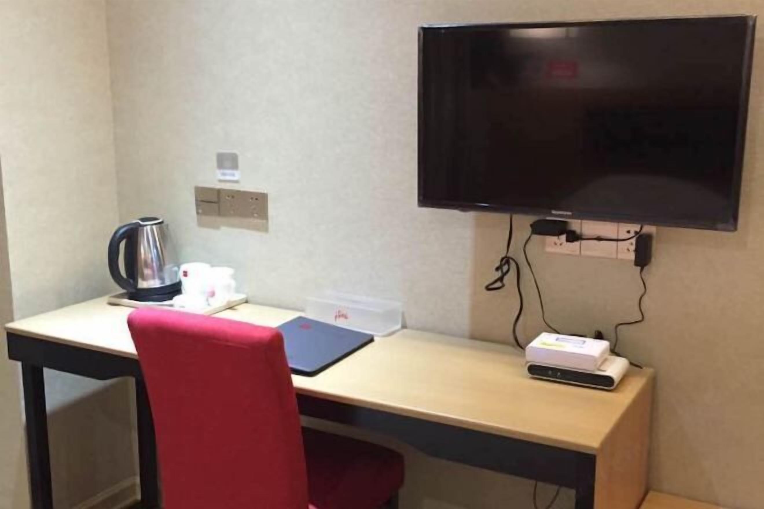 A hotel room with a desk, a red chair, a television mounted on the wall, a kettle, and a power strip with a device plugged in.
