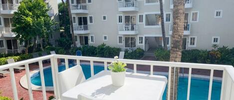 A hotel balcony with a pool, white furniture, and a view of the building's exterior.