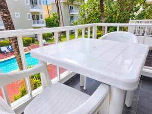 Une table et des chaises blanches, placées à l’extérieur sur un balcon, offrent une vue sur une piscine et un bâtiment situé au loin.
