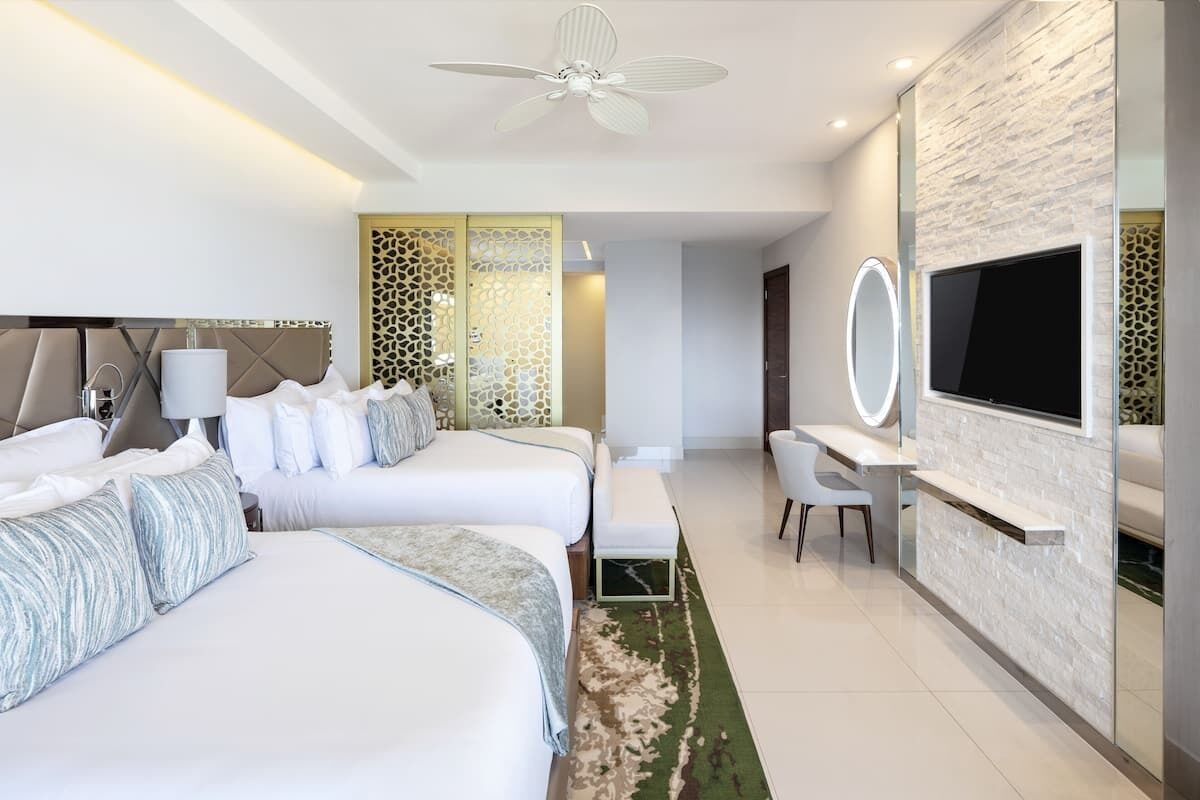 Quarto de hotel moderno com duas camas, uma escrivaninha, uma TV e um ventilador de teto.