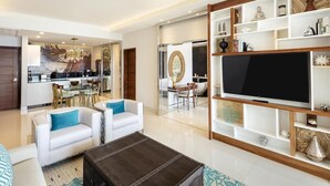 Un soggiorno moderno con una grande TV a schermo piatto, divani bianchi con cuscini blu, un poggiapiedi in pelle scura e un mobile TV in legno con oggetti decorativi.