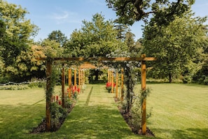 Puinen pergola, jonka ympärillä kasvaa köynnöskasveja ja kukkia, puutarhamaisessa ympäristössä.