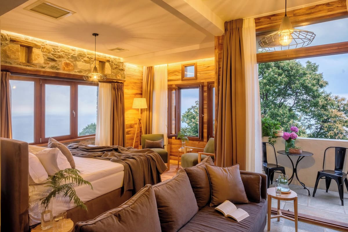 A cozy room with a bed, a sofa, a table with chairs, and a balcony with a view of trees.