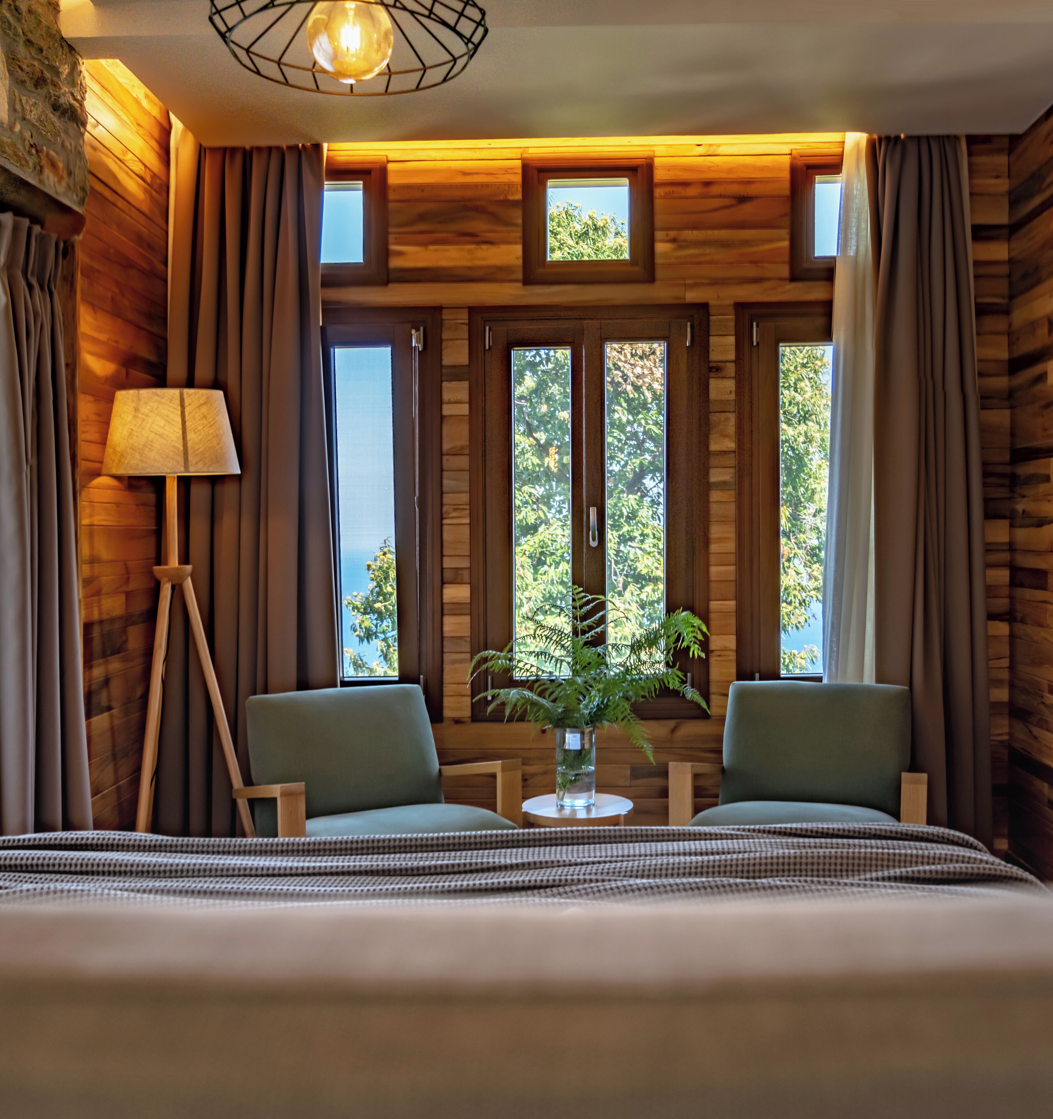 A wooden room with two chairs, a bed, a lamp, and a window with a view of trees.