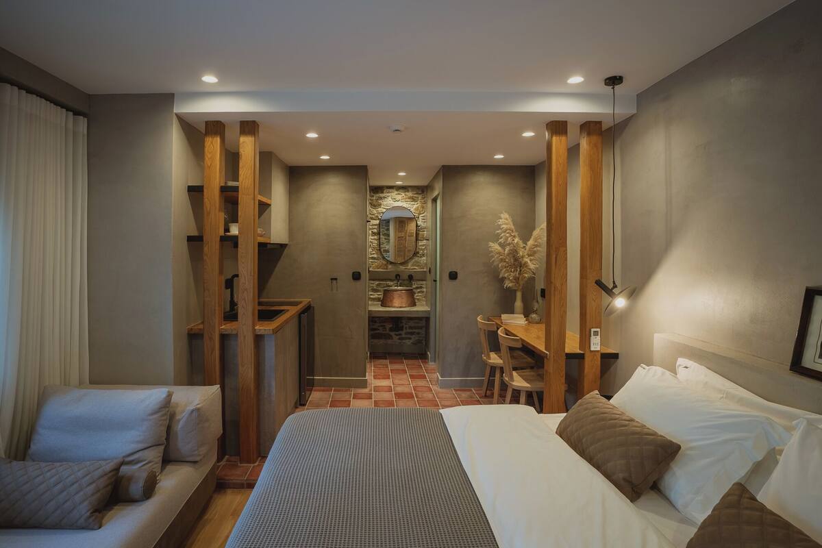 A modern bedroom with a bed, a desk, and a chair. There is a small kitchen area with a sink and a stove. The room has wooden beams and a stone wall.