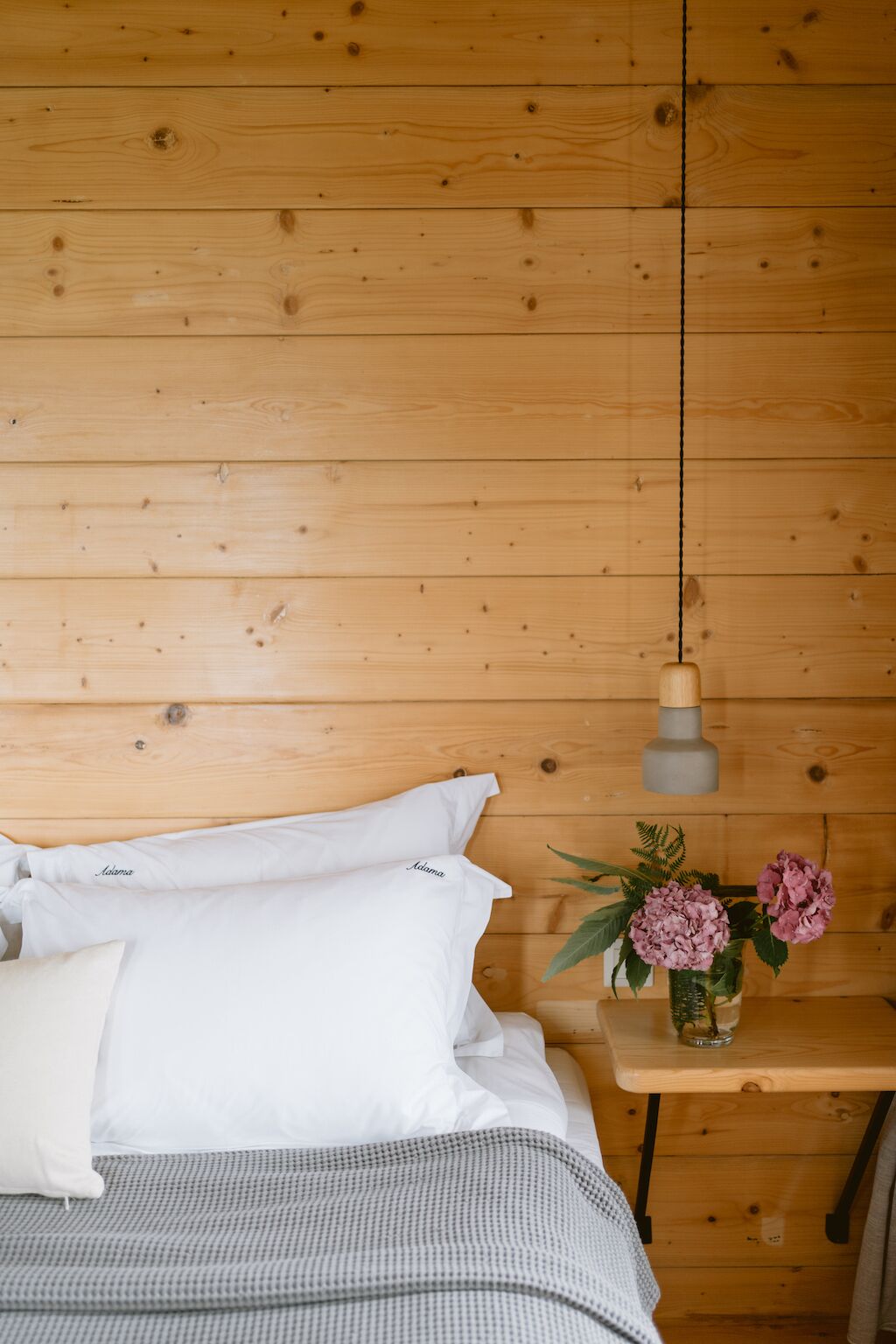 Habitación con paneles de madera, una cama, almohadas blancas, una manta gris a cuadros, una mesita de noche de madera con un jarrón de flores rosas y una lámpara colgante.