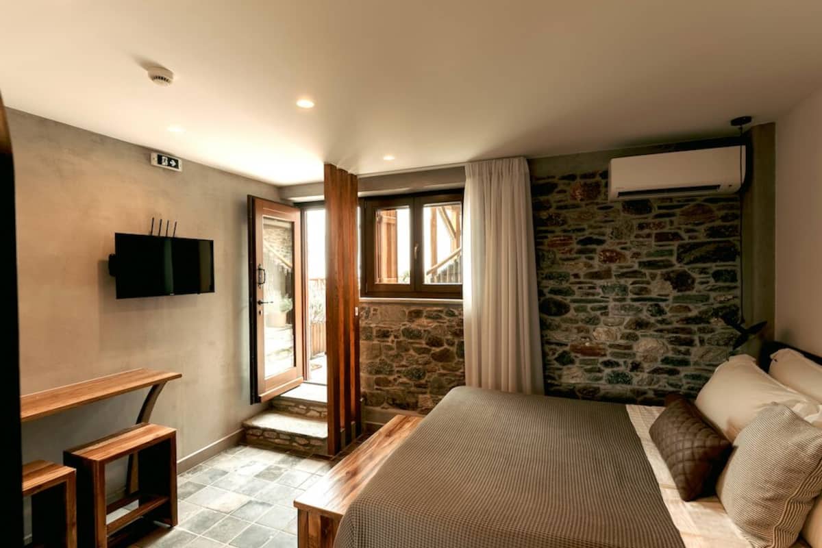 A bedroom with a stone wall, a bed with pillows, a wooden bench, and a TV on the wall.