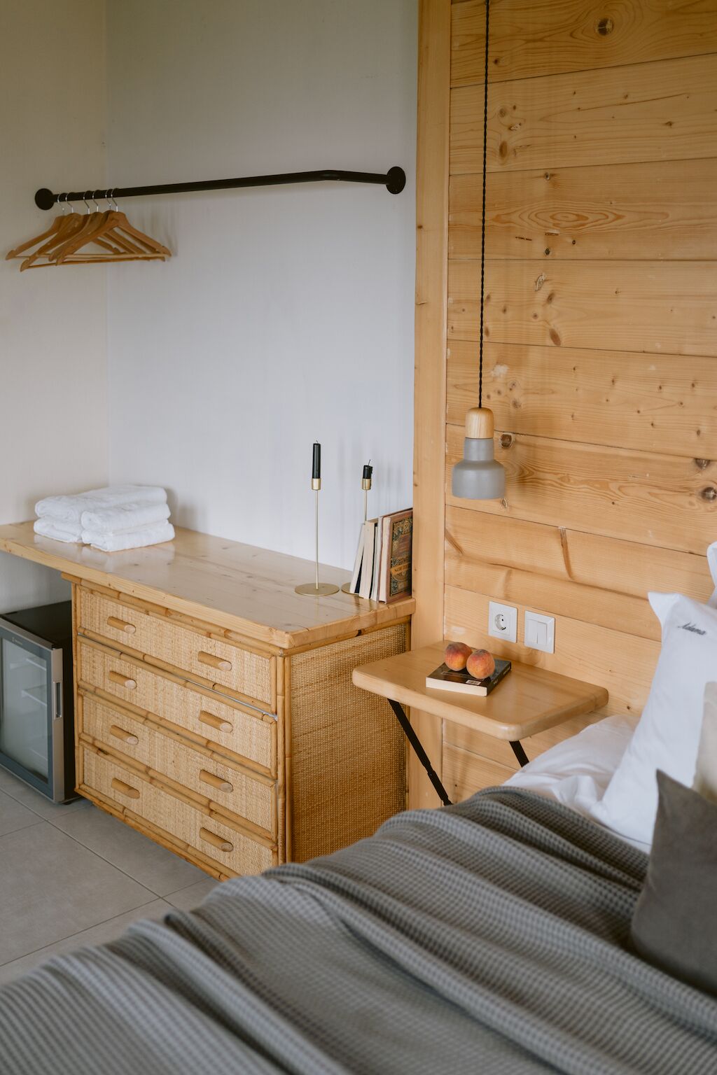 Quarto com cabeceira de madeira, mesa de cabeceira com luminária, cama com manta listrada e pequena cômoda de madeira com gavetas.