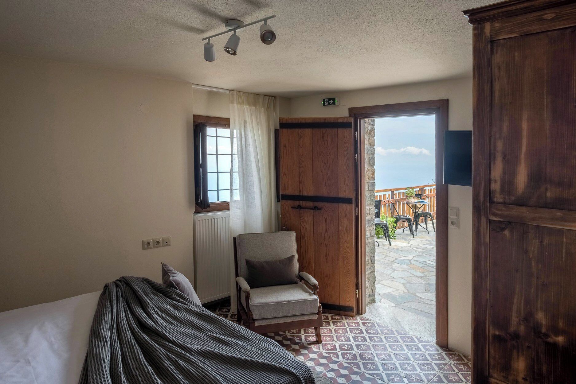 A hotel room with a bed, a chair, a wooden wardrobe, a window with curtains, and a view of an outdoor patio.