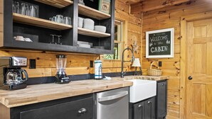 A kitchen with wooden cabinets, a coffee maker, a blender, and a sink. There is a welcome sign on the wall and a basket on the counter. - Cozy Mountain Cabin w/ Hot Tub Close to Gatlinburg (Sevierville)
