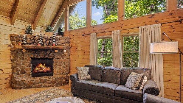 A cozy cabin interior with a stone fireplace, a leather sofa, and a large window overlooking greenery. - Cozy Mountain Cabin w/ Hot Tub Close to Gatlinburg (Sevierville)