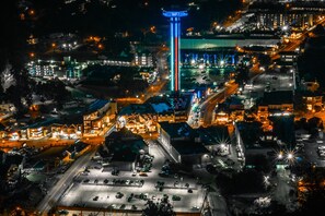 An aerial view of a cityscape at night with illuminated buildings, a prominent tower, and a parking area.
