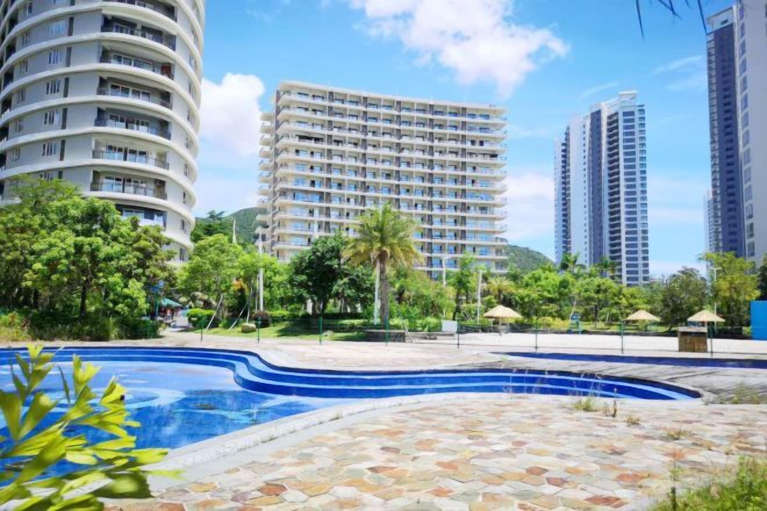 Una piscina con borde de baldosas, rodeada de vegetación y edificios altos.