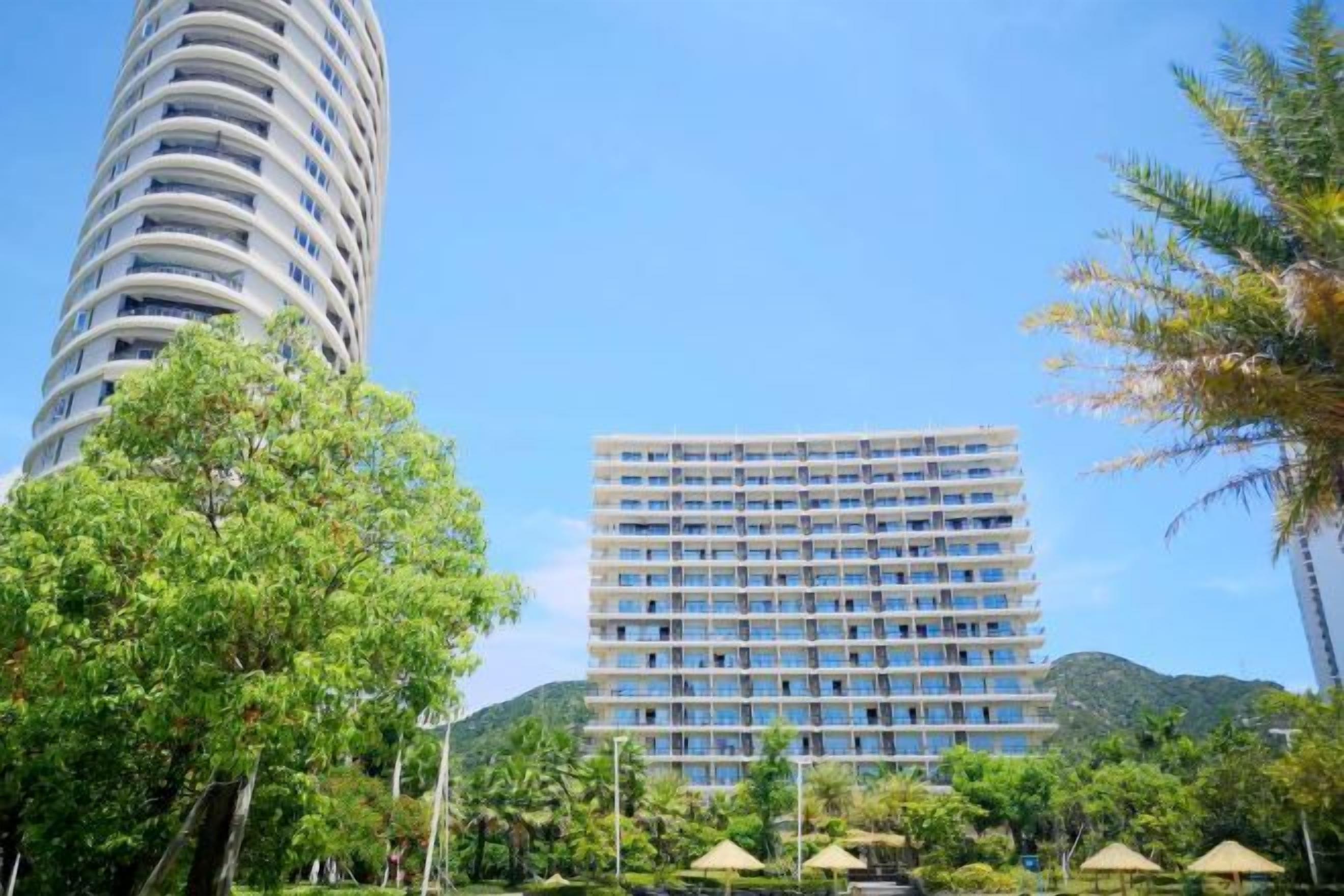 Un hotel moderno con una torre cilíndrica y un edificio rectangular, rodeado de vegetación y un cielo despejado.