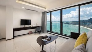 A modern hotel room with a large window, a flat-screen TV, a sofa, a coffee table, and a desk with a chair.