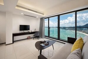 A modern hotel room with a large window, a flat-screen TV, a sofa, a coffee table, and a desk with a chair.