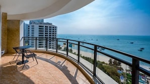 A balcony with a curved railing overlooking a beach and ocean, a table, and a chair.
