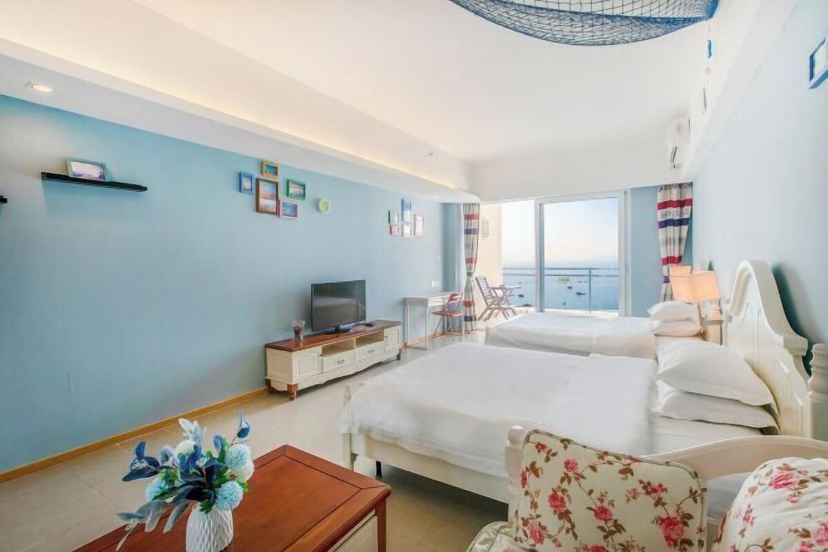 A hotel room with two beds, a TV, a desk, a chair, and a view of the sea.