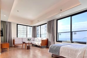 A modern hotel room with a large bed, a sofa, a coffee table, and a view of the city through large windows. - Yimi Yangguang Seaview Holiday Apartment (Huidong)