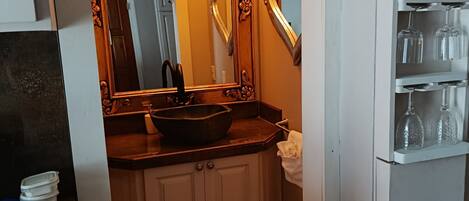 A bathroom with a vanity, a large mirror with lights, a sink, and a shelf with glassware.