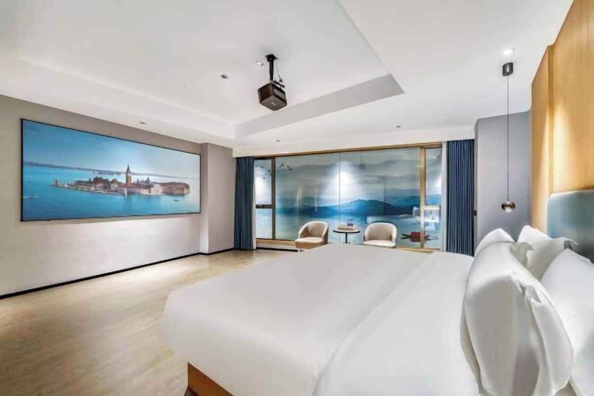 Een moderne hotelkamer met een groot bed, een schilderij van een kustplaats, een balkon met uitzicht en een projector aan het plafond.