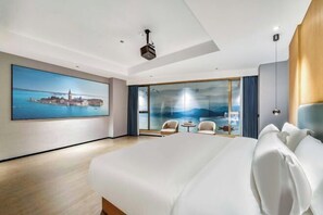 Et moderne hotelværelse med en stor seng, et maleri af en kystby, en balkon med udsigt og en projektor i loftet.