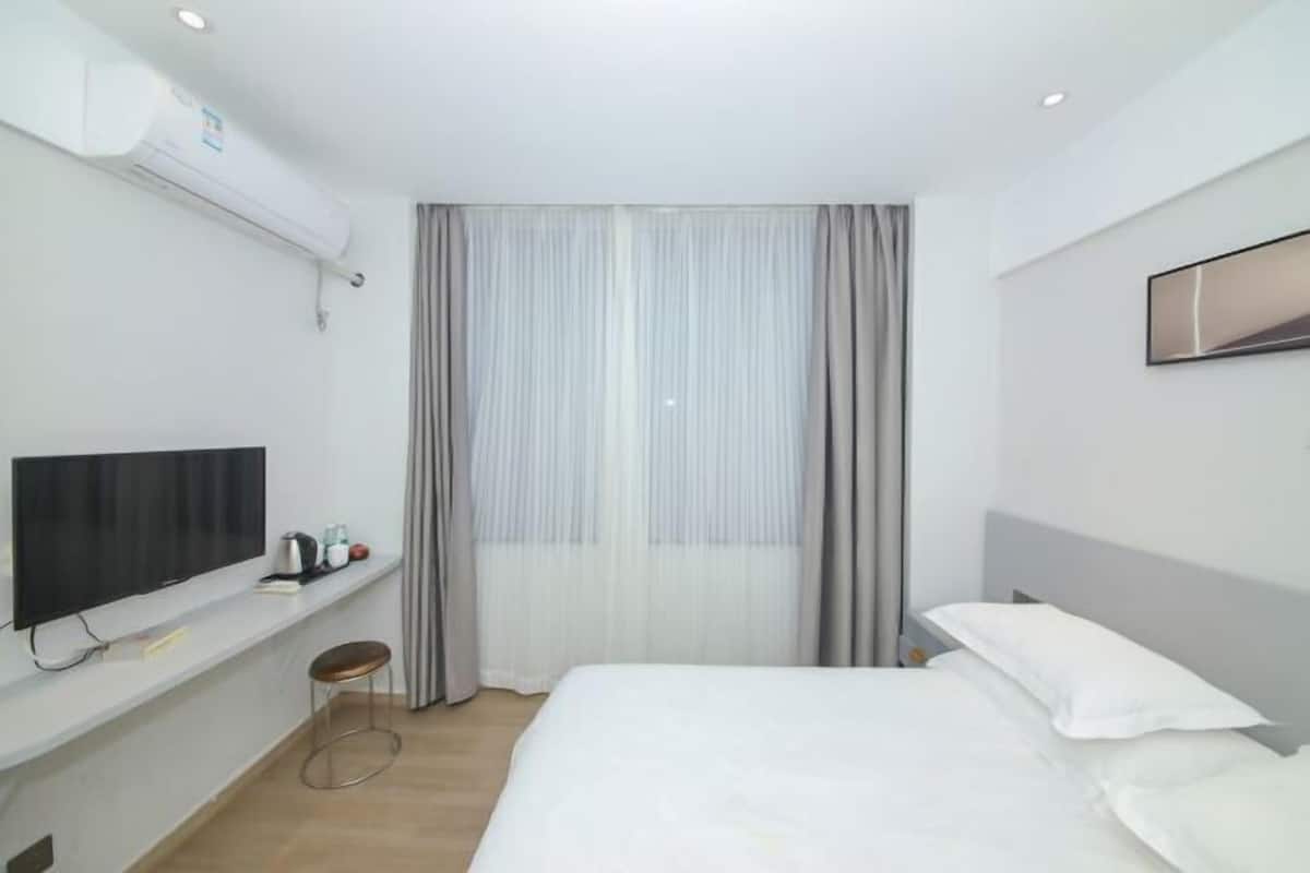 Ein Hotelzimmer mit einem Bett, einem Fernseher, einem kleinen Hocker, einem Fenster mit Vorhängen und einer Wandklimaanlage.