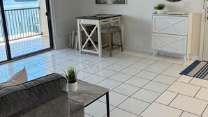 A room with a ceiling fan, a desk with drawers, a chair, a sofa, a small table with a plant, a round mirror, and a view of a body of water through a sliding door.