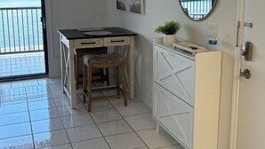 A room with a tiled floor, a desk with a chair, a white cabinet, a mirror, and a view of a body of water through a door.