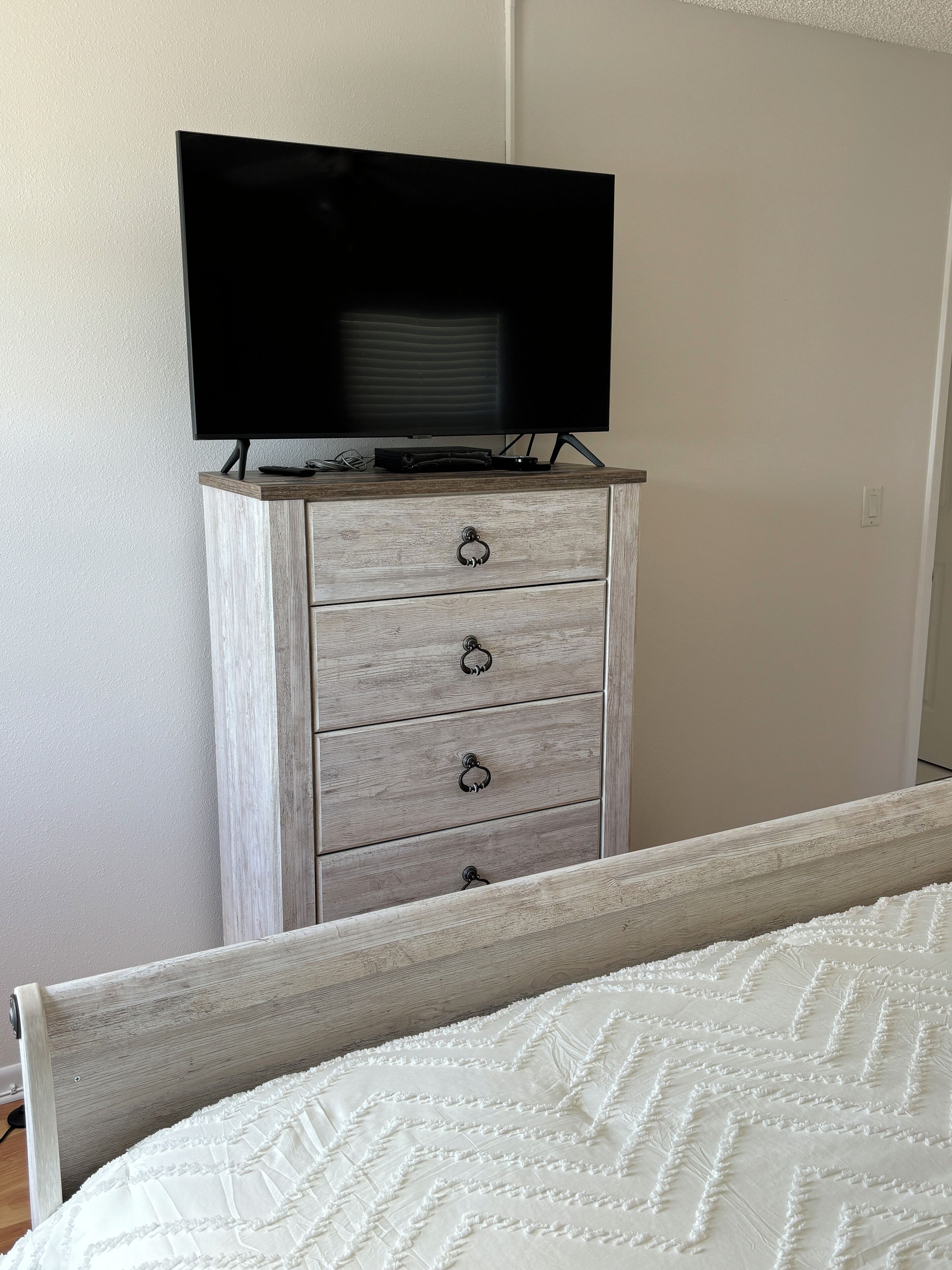 Une chambre à coucher avec un lit, une commode sur laquelle est posée une télévision, et un mur avec un interrupteur.