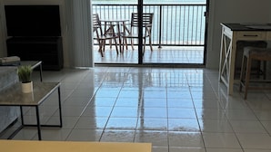 A spacious living room with a large flat-screen TV, a ceiling fan, and a balcony with a view of a body of water.