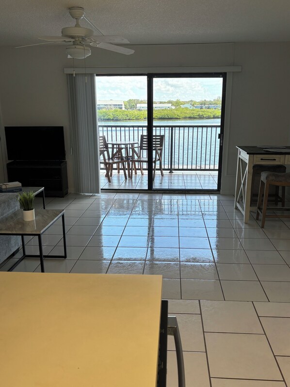 A spacious living room with a large flat-screen TV, a ceiling fan, and a balcony with a view of a body of water.