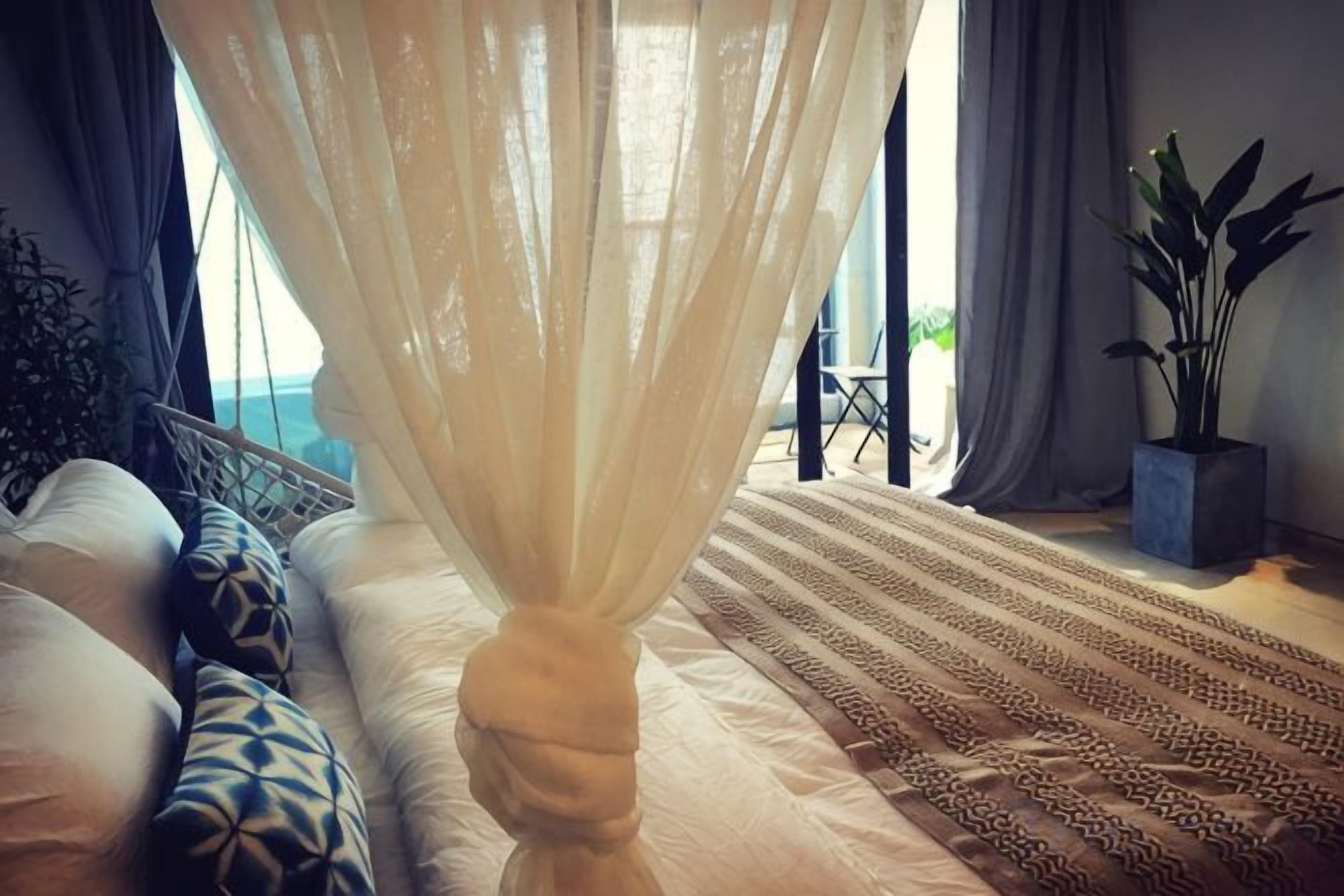 Une chambre avec un lit, un fauteuil suspendu, une plante en pot et une vue sur l’extérieur.