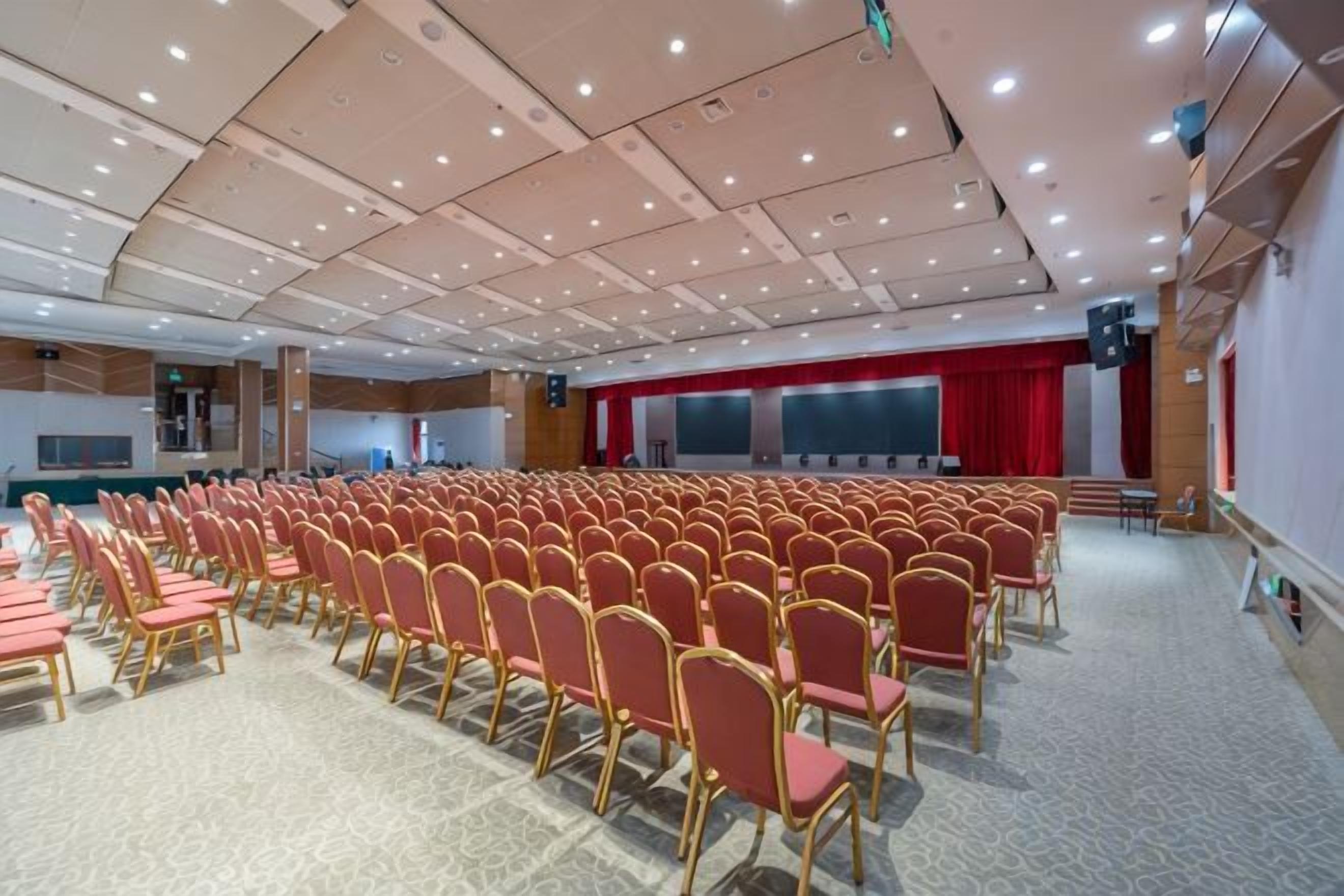 A conference room with rows of chairs, a stage with a red curtain, and a carpeted floor.