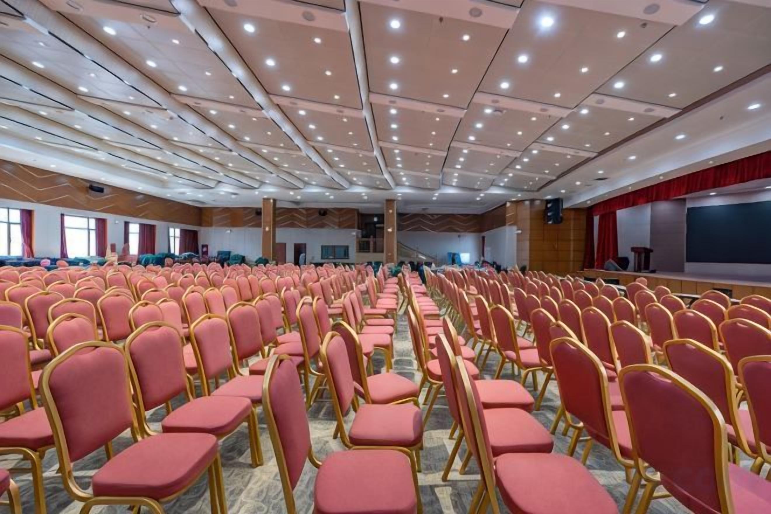 Un amplio salón de conferencias con filas de sillas, un escenario y una pantalla de proyección.